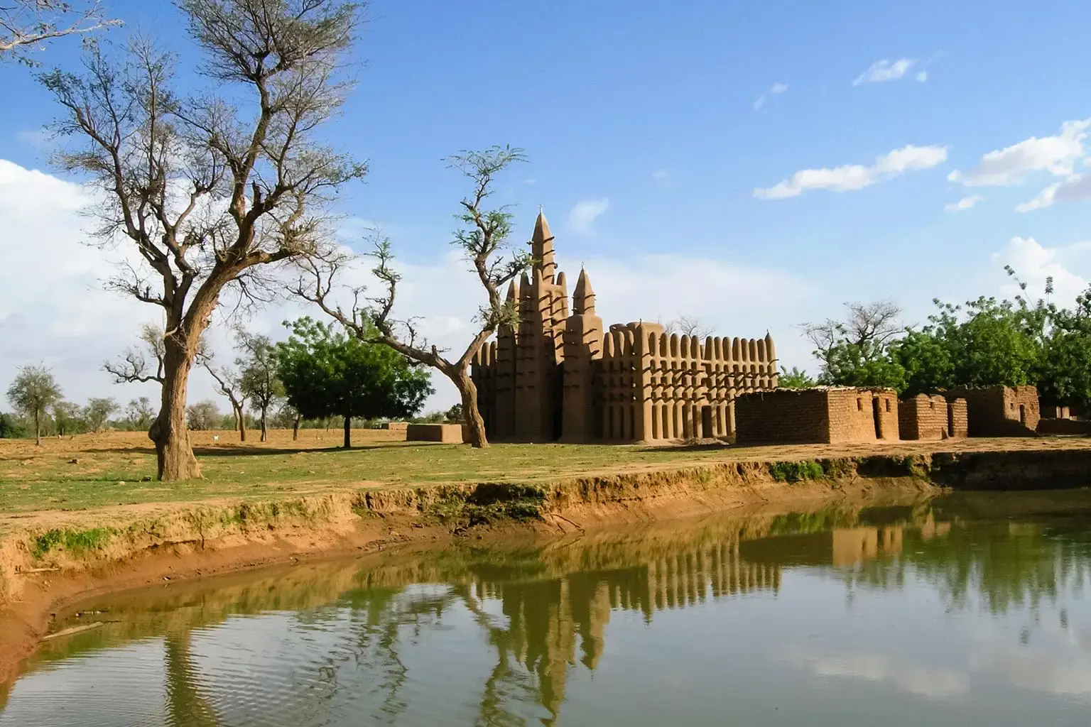 Mosquée en banco aux tours crénelées se reflétant dans une étendue d'eau, entourée d'arbres