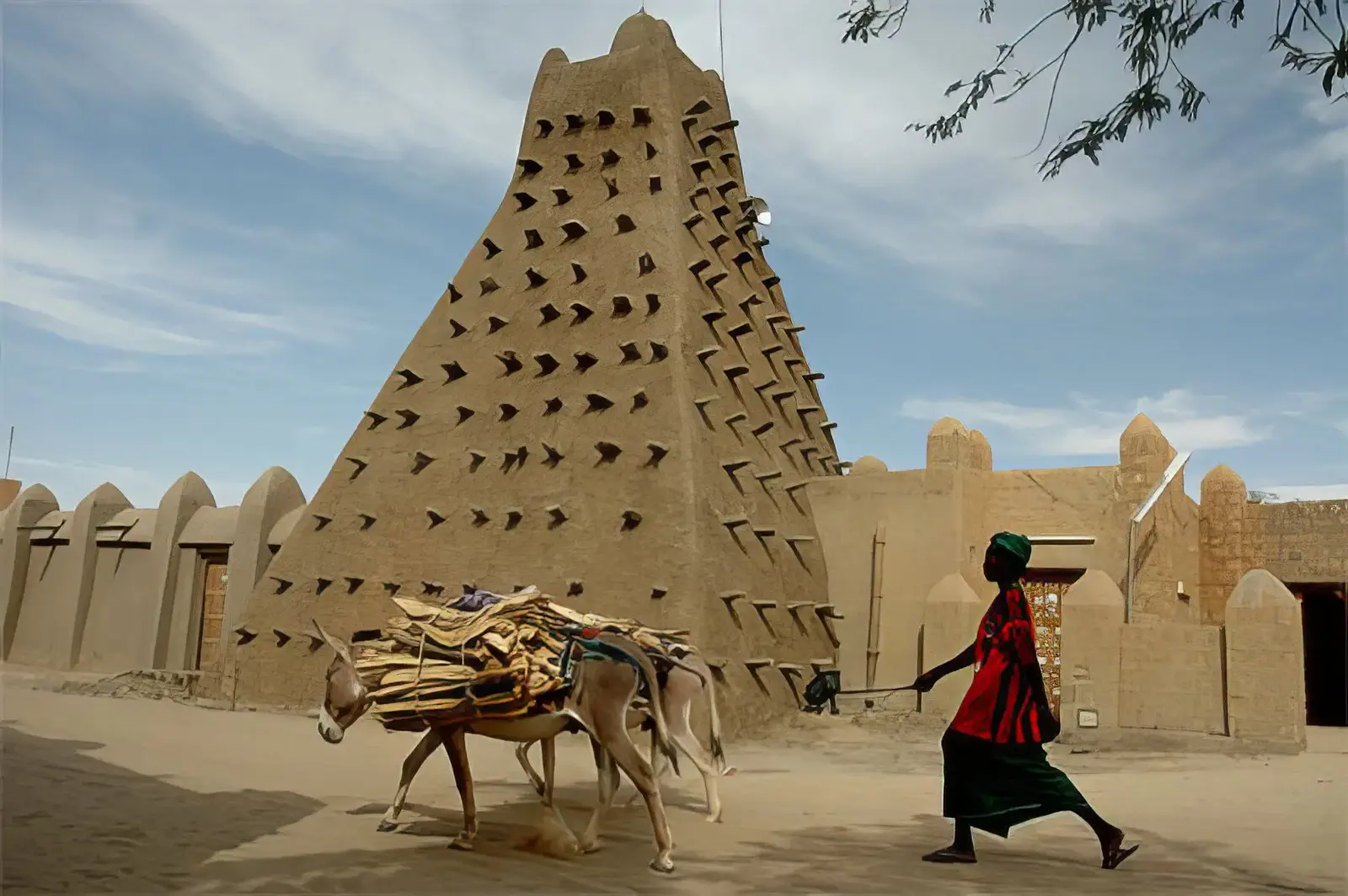 Minaret pyramidal en banco hérissé de poutres ; une passante et un âne chargé de bois au premier plan